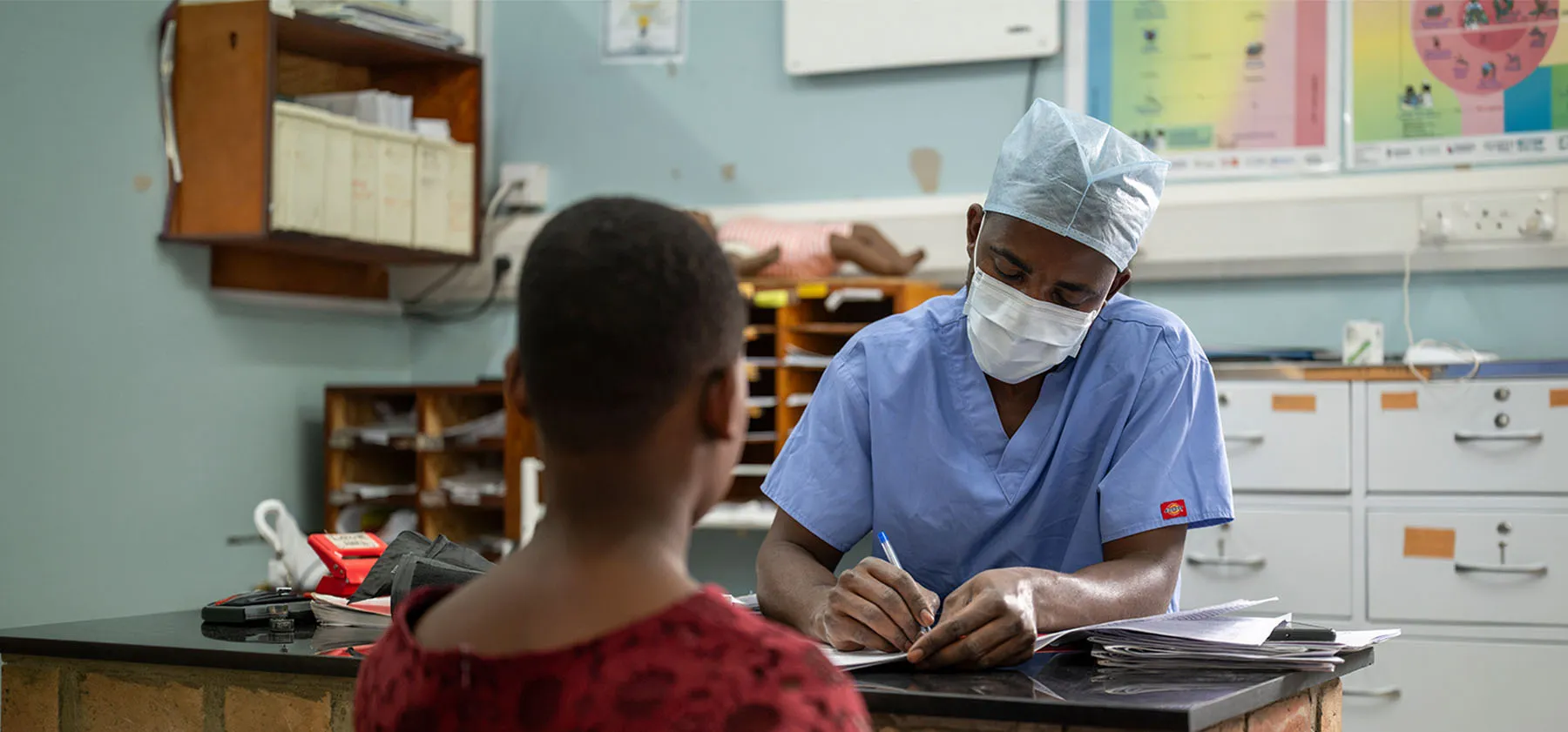 A clinician in a surgical cap and mask writes notes while speaking with a patient.