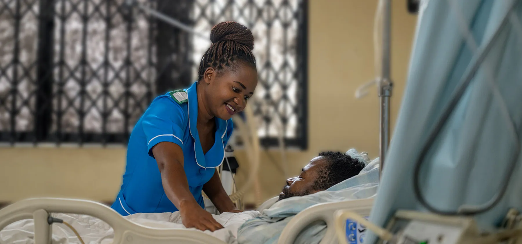 A smiling nurse in blue scrubs leans over a patient resting in a hospital bed.