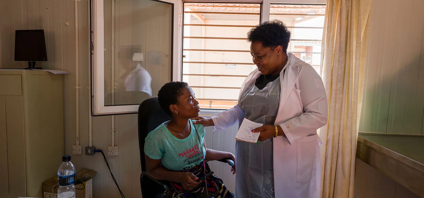 A clinician in a white coat places a reassuring hand on the shoulder of a young patient in an exam room.