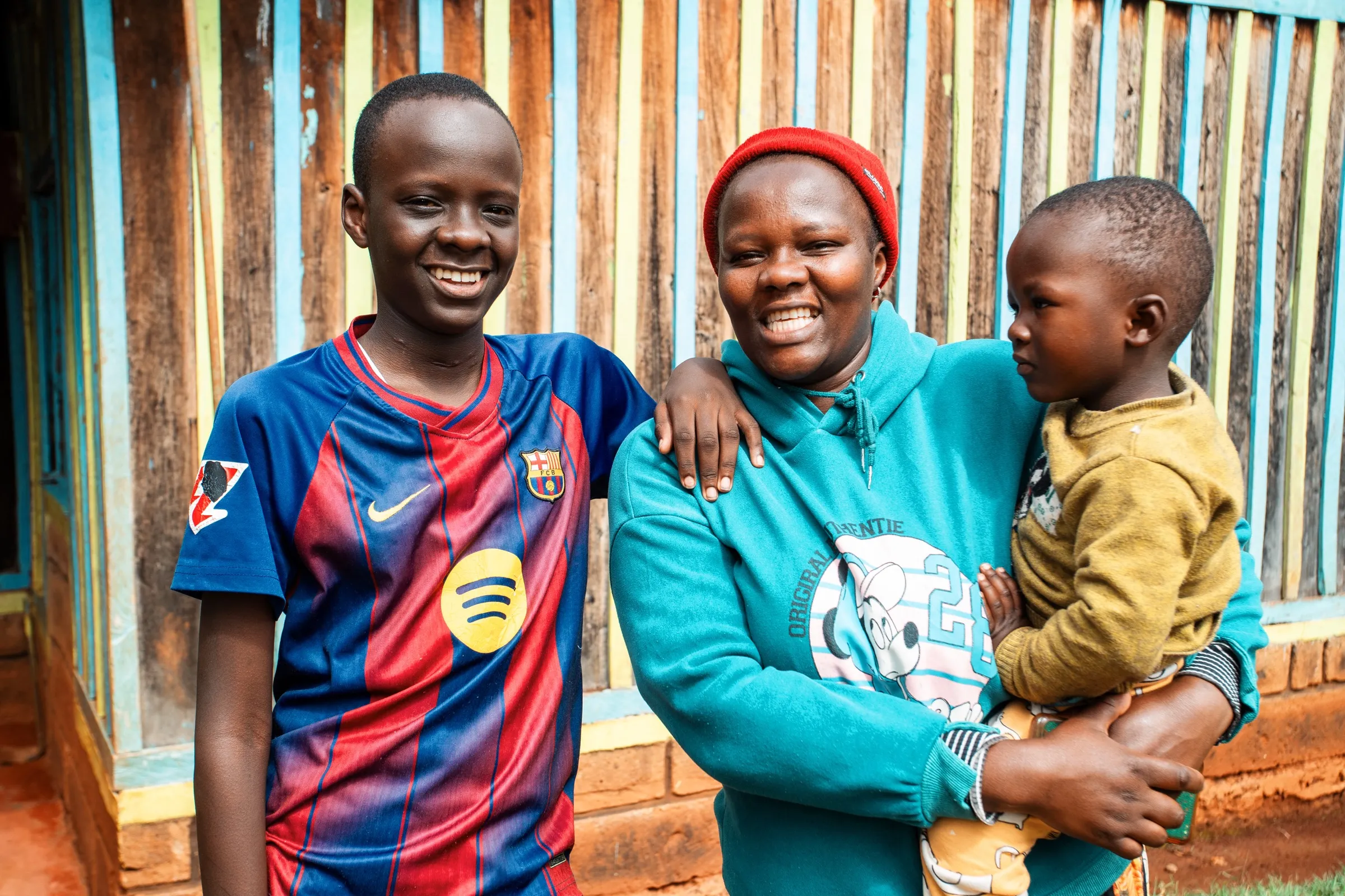 Winston stands smiling outside his home in Kenya beside his mother, who holds a young child on her hip.